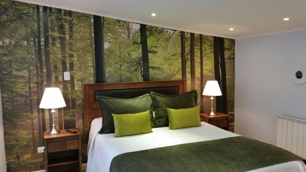 Bedroom with a forest mural behind a wooden headboard, white bedding, and green cushions; two bedside tables with lamps flank the bed.