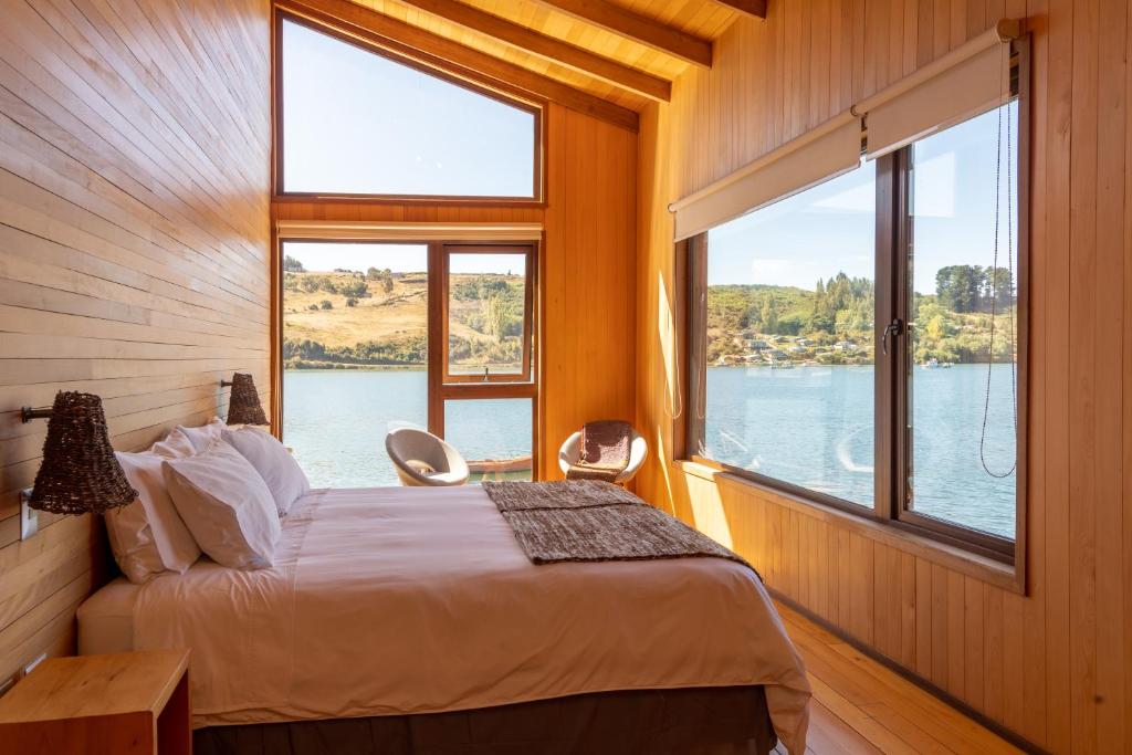 Wood-paneled lakeside bedroom with a white-bed, two chairs by the window, and a view of a calm lake and hills outside.