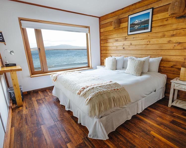 Cozy bedroom with ocean view: white bed with pillows and cream throws, wood-paneled wall, and a large window showing the sea.