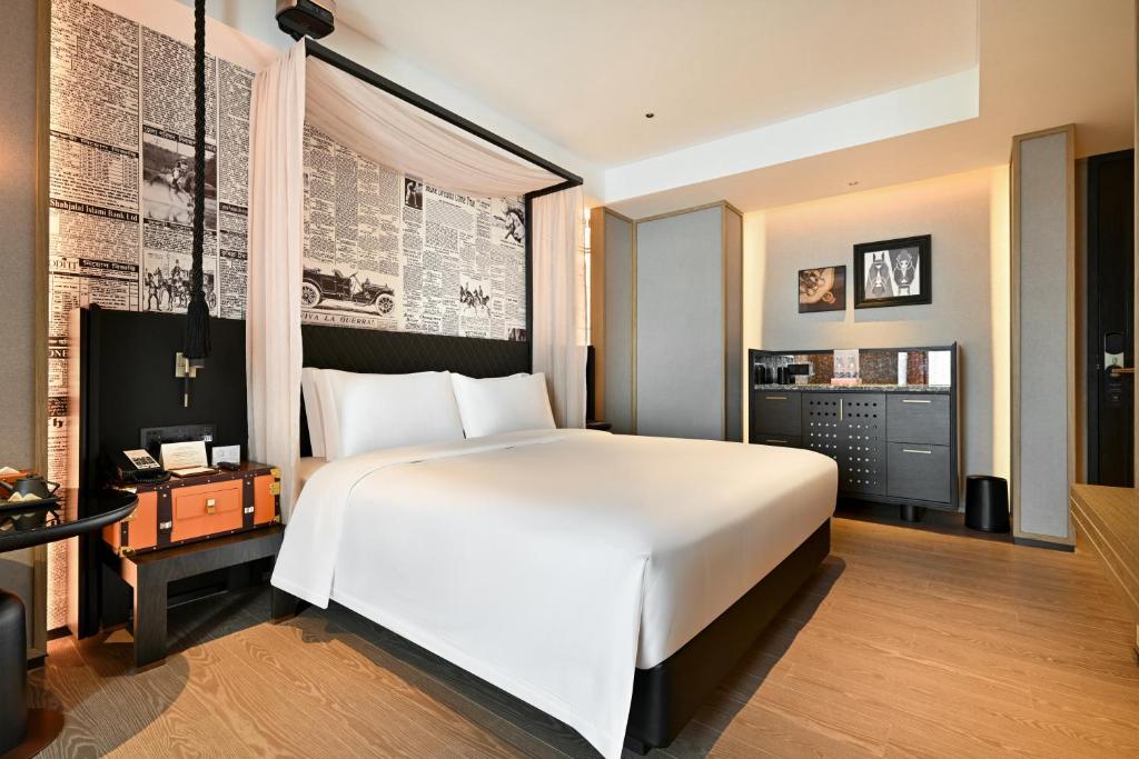 Modern hotel room with a white canopy bed, newspaper-patterned headboard wall, and a vintage orange-trunk nightstand.