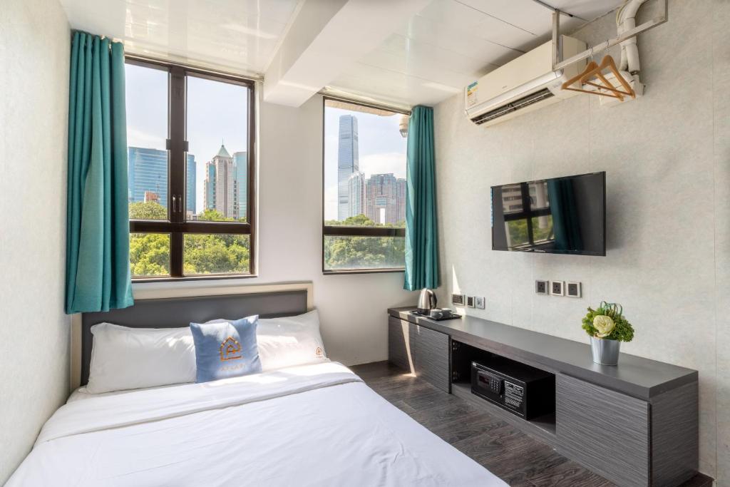 Hotel room with a white bed, teal curtains, and a wall-mounted TV; city skyline visible through two large windows.