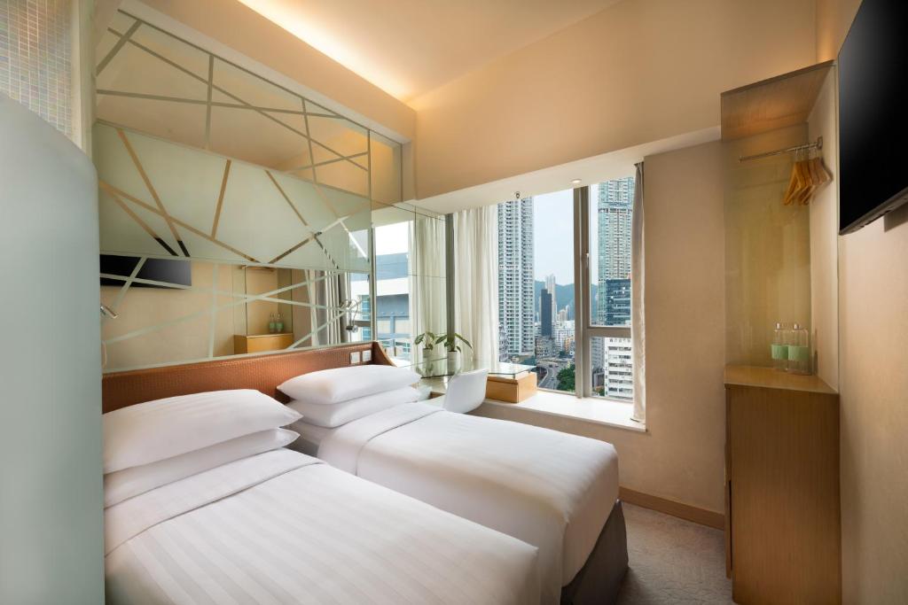 Two twin beds with white linens in a bright hotel room, a glass partition with geometric lines, and a city view through a large window.