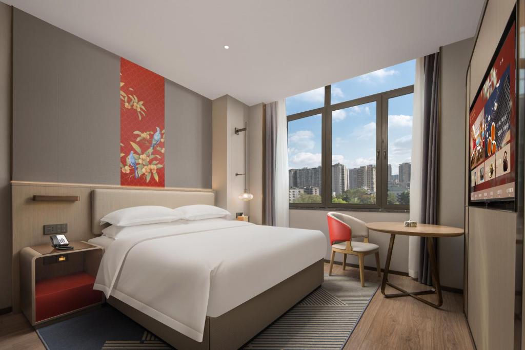 Hotel room with a king bed, round table and two chairs by a large window overlooking a cityscape, plus wall art above the bed.