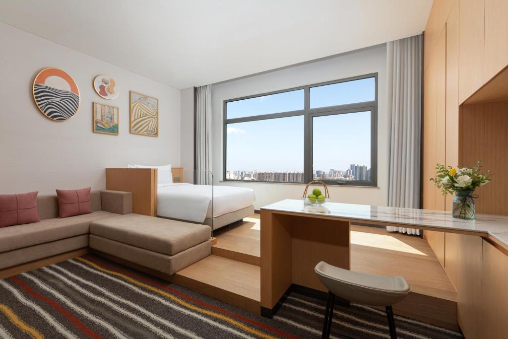 Hotel suite with a beige sectional sofa, elevated bed, and a large window showing the city skyline beneath a clear blue sky, in a bright room with wooden furniture.