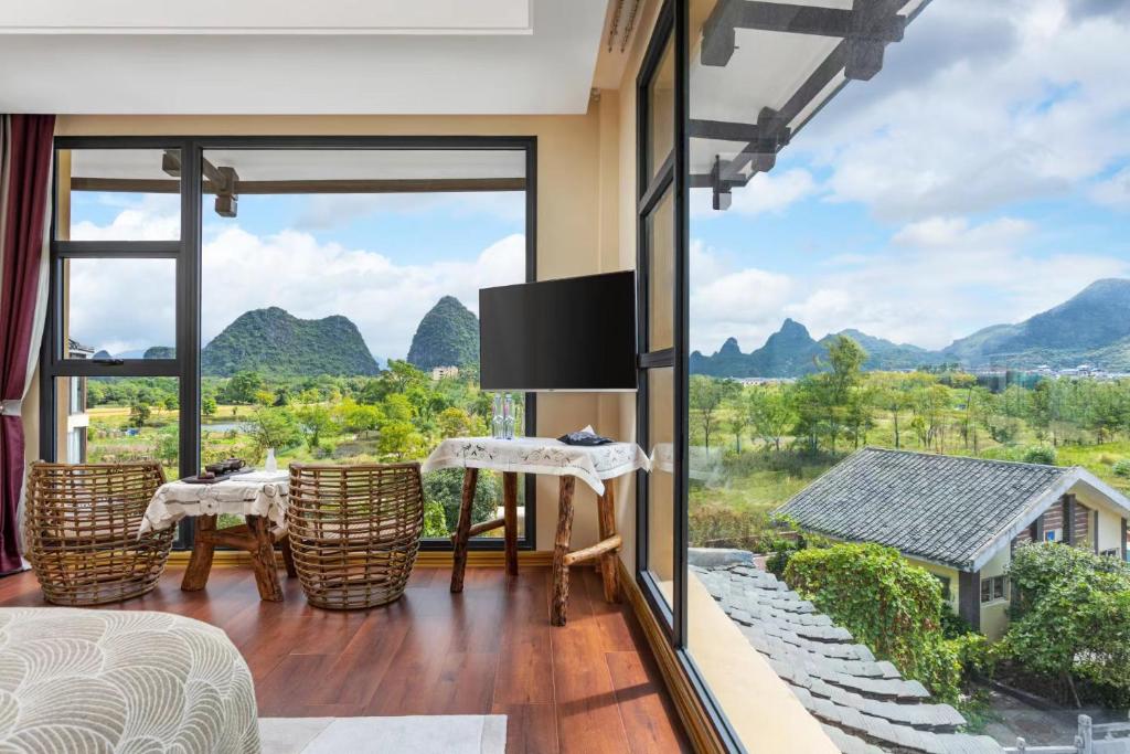 Bright hotel room with floor-to-ceiling windows framing a mountain landscape; wicker chairs sit around a small table on a wooden floor