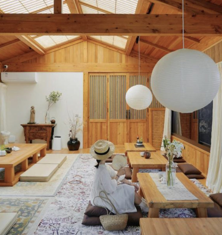 Warm wooden interior with low tables, floor cushions, and large white paper lanterns; a person wearing a hat sits at a table.