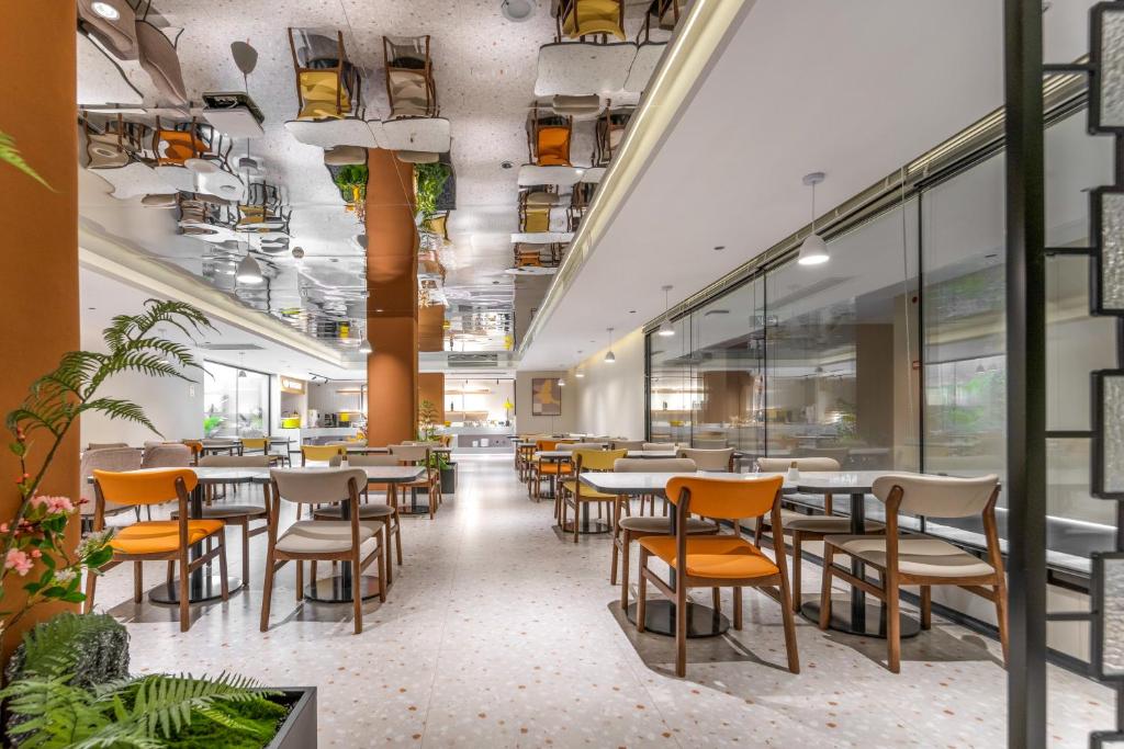 Spacious cafe with orange and beige chairs around round and rectangular tables, bright lighting, and a mirrored ceiling reflection down the center aisle.