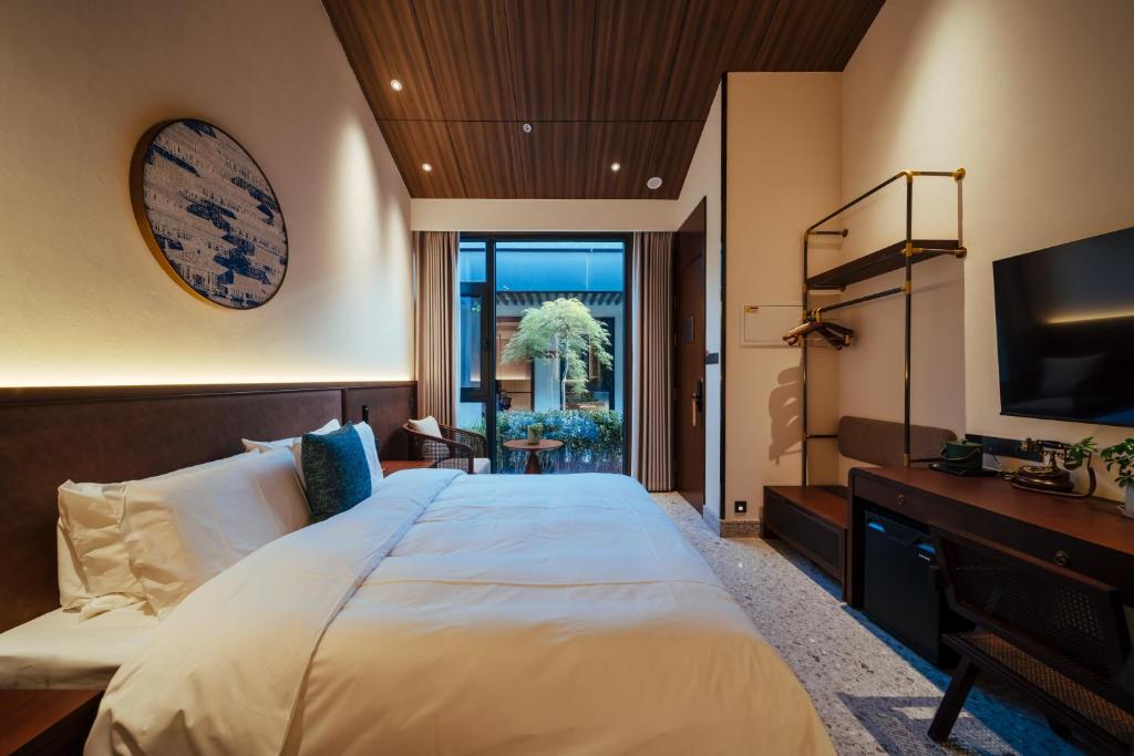 Modern hotel room with a large white-lynens bed, a seating area, and a wall-mounted TV opposite a desk and chair; glass doors open to a small outdoor area.