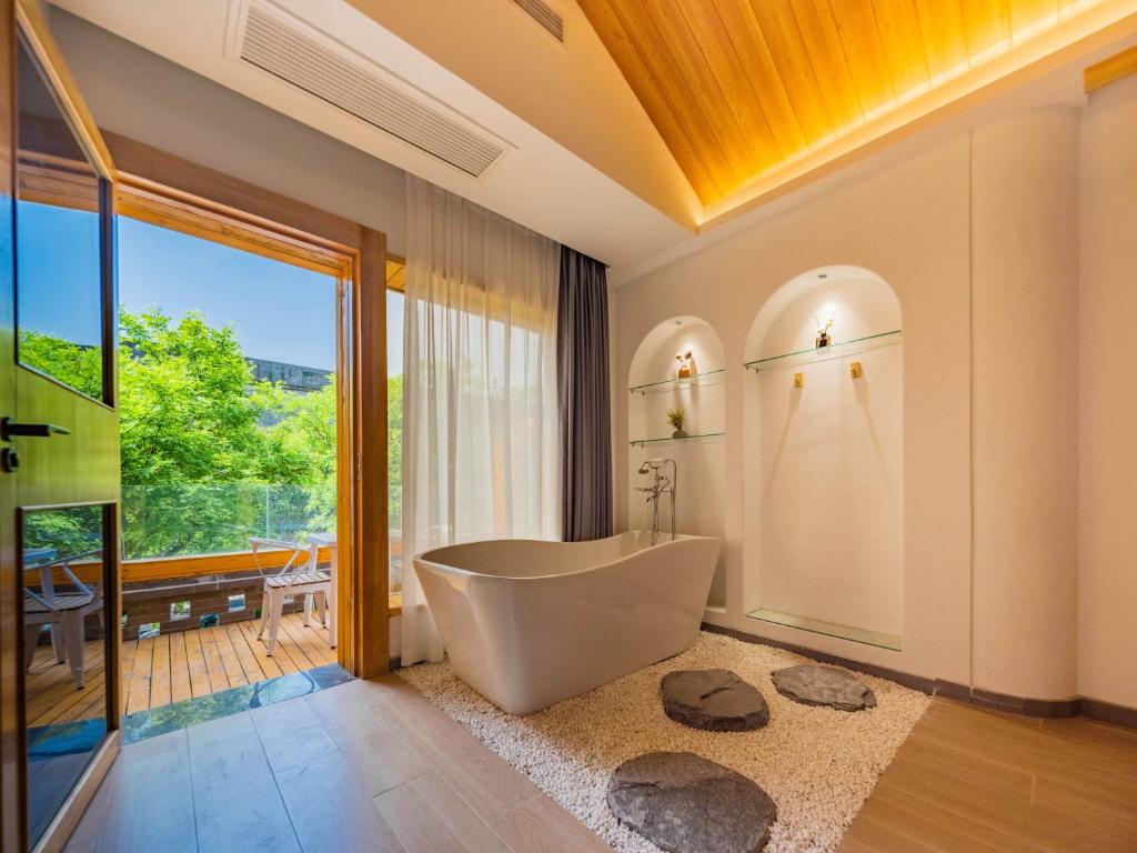 Modern bathroom with a white freestanding tub, pebble floor, and built-in glass shelves; door opens to a wooden balcony with greenery outside.