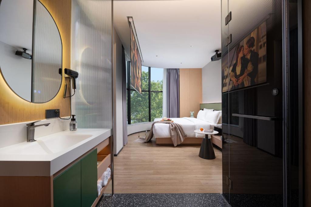 Hotel suite with a sink and round mirror on the left, leading to a bedroom with a large window and greenery outside.