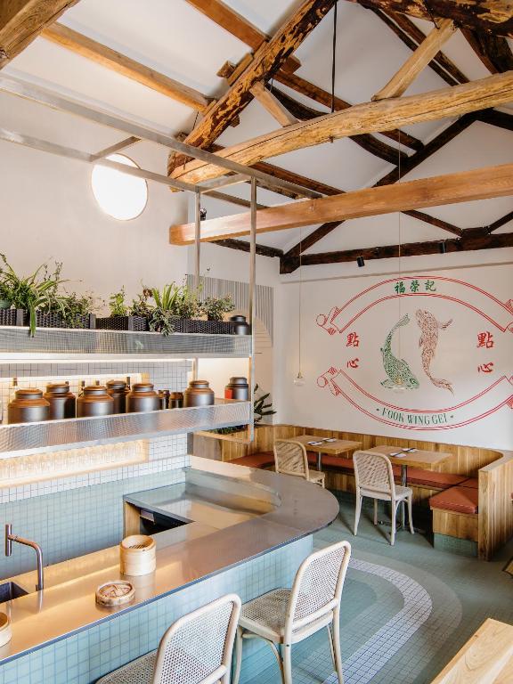 Cafe interior with curved stainless counter, jars on shelves, potted plants, and wooden beam ceiling above a seating area against a wall mural with koi fish and a circular logo