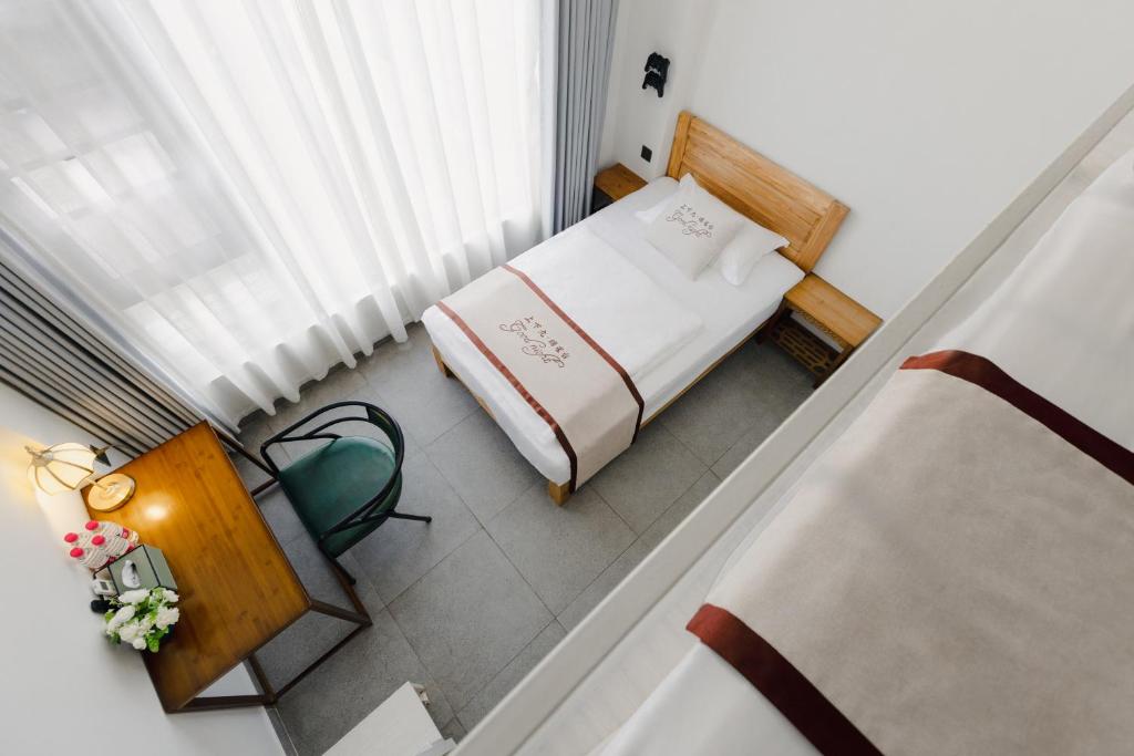 Small hotel room with a single wooden bed, white bedding with a brown stripe, a matching nightstand, desk, and a green chair beside a large window with sheer white curtains.