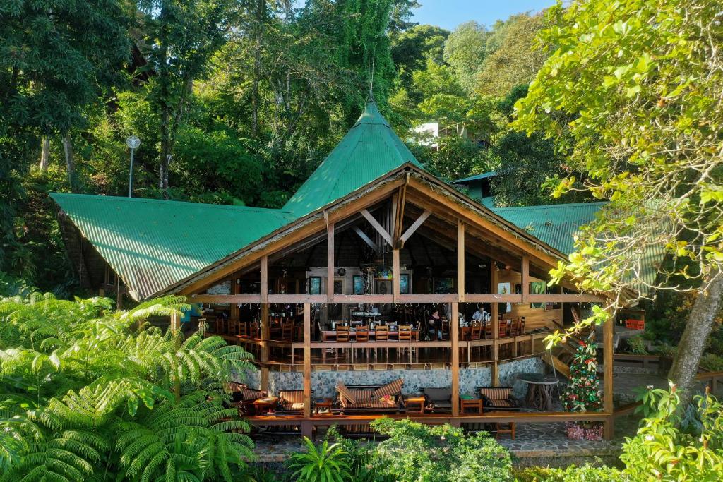 Open-air wooden restaurant with a green roof set in a tropical forest; diners visible inside on a covered deck.
