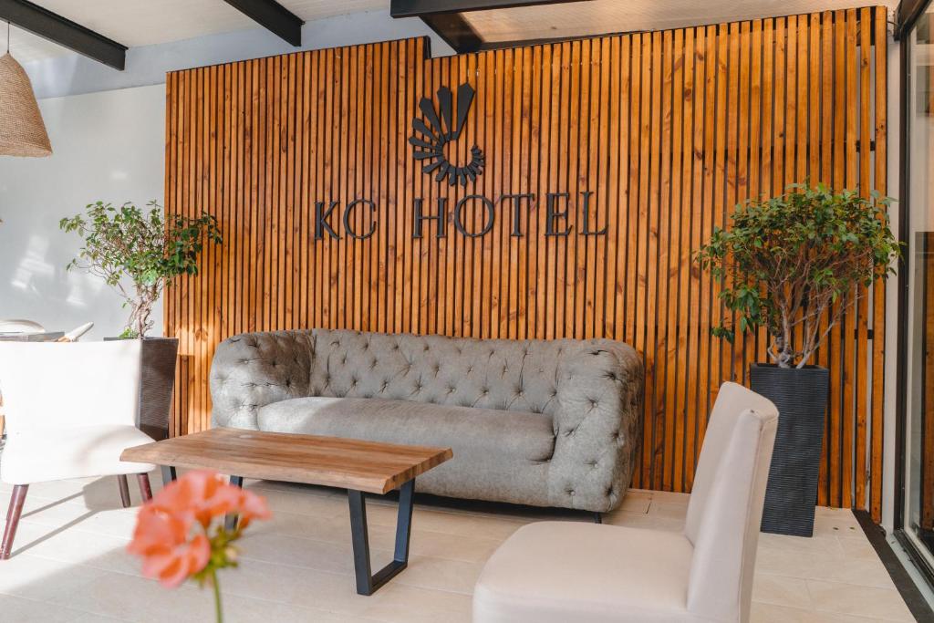 Wood-paneled wall with KC HOTEL logo behind a tufted gray sofa and a wooden coffee table in a bright hotel lobby with potted trees on both sides.