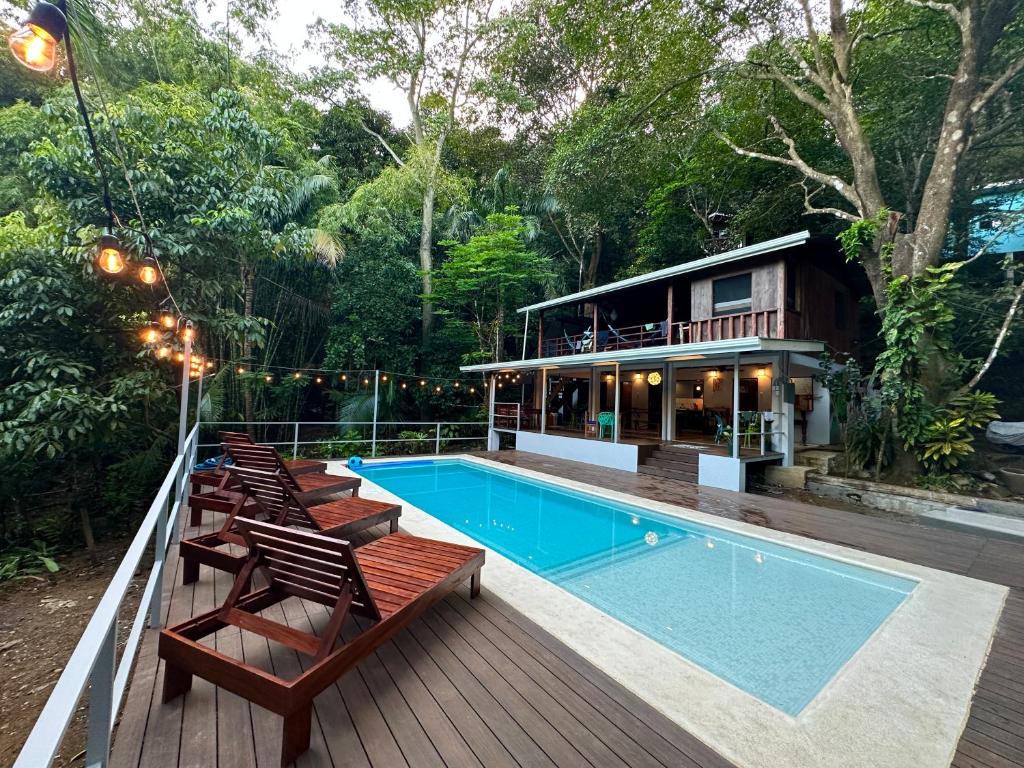 Outdoor pool area with four wooden lounge chairs along a deck, string lights, and a two-story wooden house surrounded by trees.