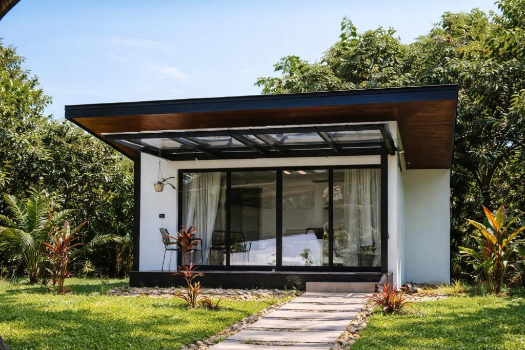 Modern glass-walled cottage with a covered patio, surrounded by greenery and a stone-pathed lawn.