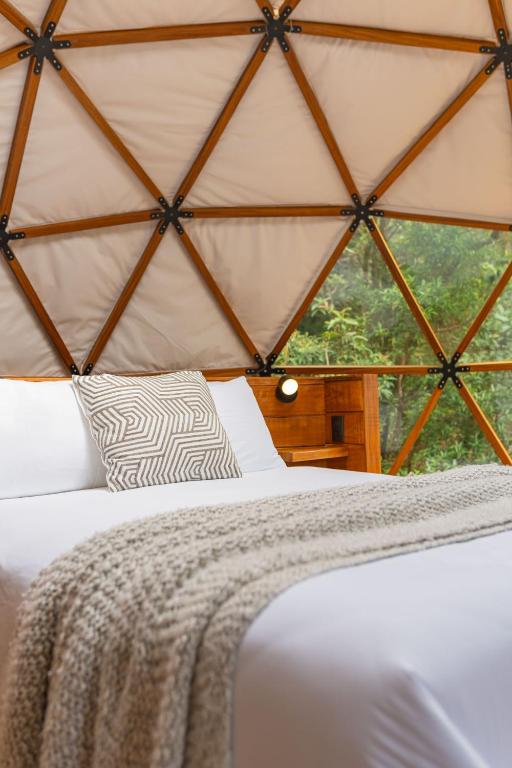 Geodesic-dome bedroom with a white bed, gray patterned pillow, and a chunky knit blanket; wooden framework and greenery visible through a side window.