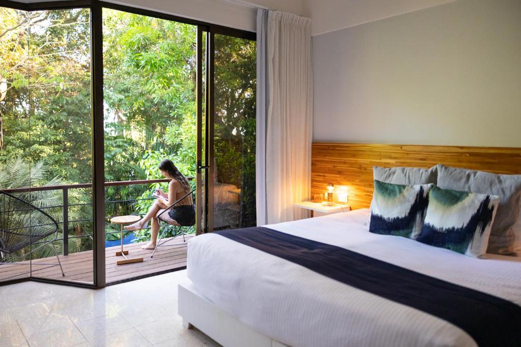 Hotel room with a sliding glass door opening to a balcony; a woman sits on a chair outside reading amid greenery outside the window.