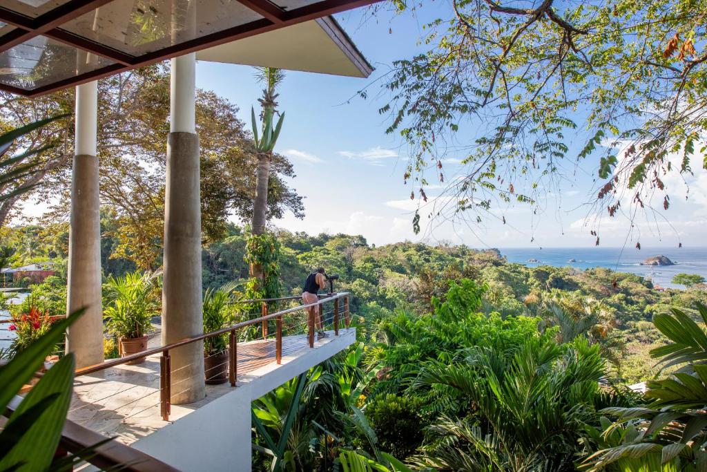 Person on a balcony with a camera overlooking lush tropical plants and a distant blue sea.