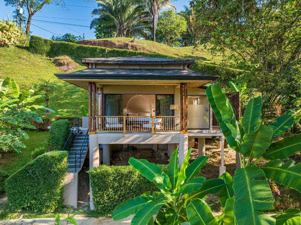 Small raised bungalow with a wooden balcony, set on a hillside among lush tropical plants and trees.