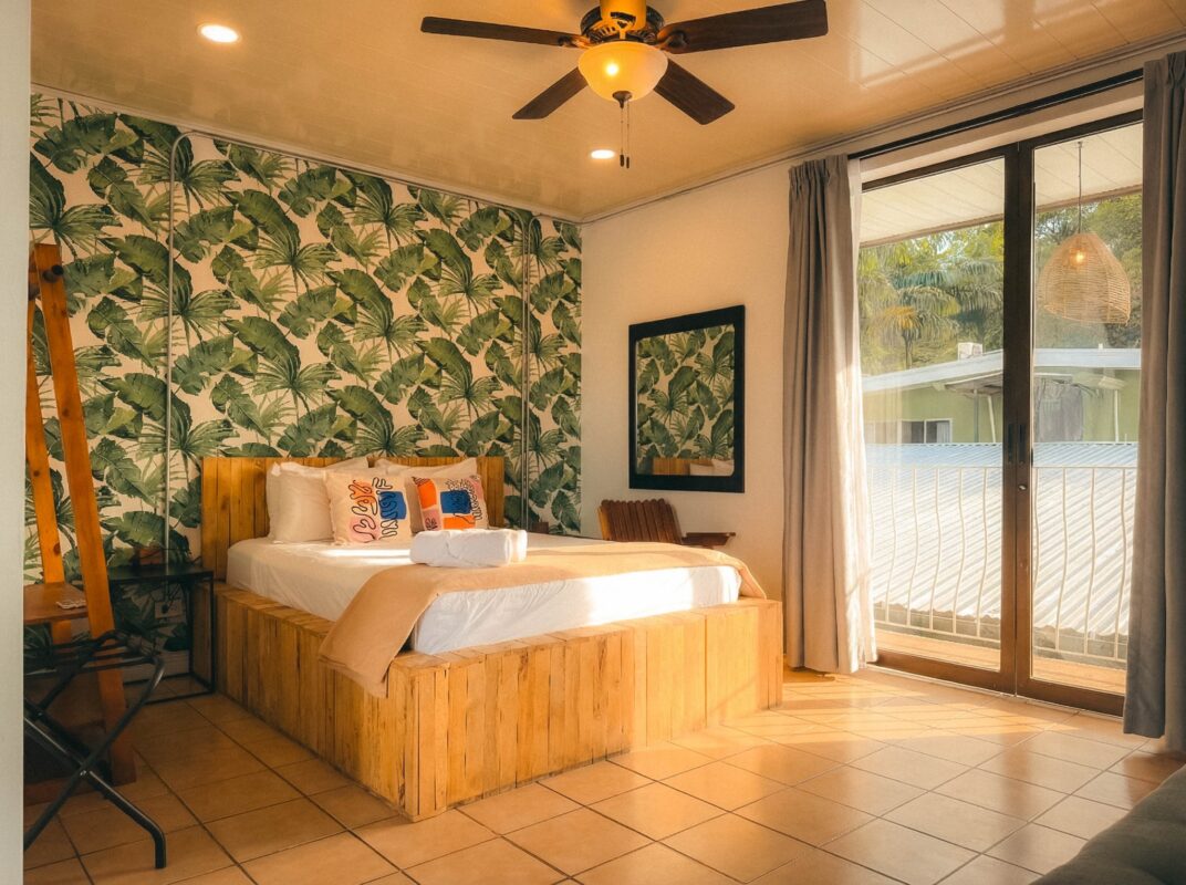 Wooden platform bed in a tropical-themed room with palm-leaf wallpaper and a rolled towel on the bed