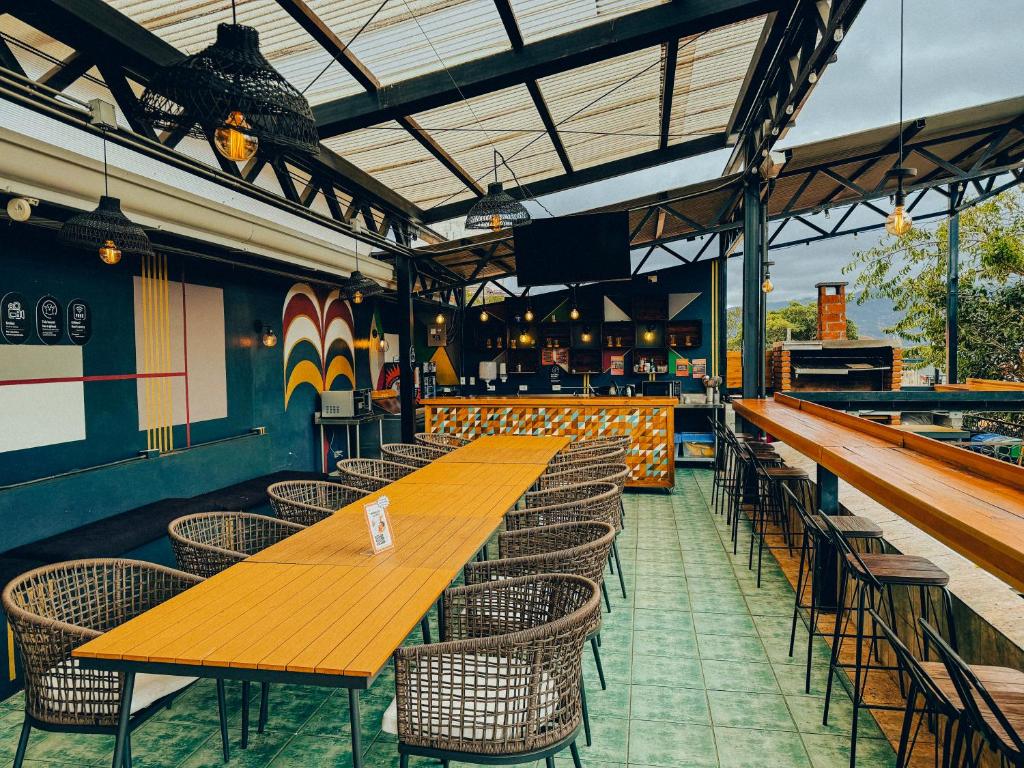 A bright outdoor bar and seating area with a long orange wooden table and wicker chairs under a glass roof, colorful mural on the wall behind.