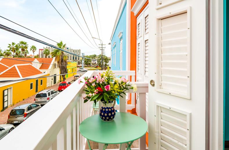 Balcony with a green table and blue patterned vase of flowers, overlooking a colorful street lined with pastel houses and parked cars under wires and a bright sky.