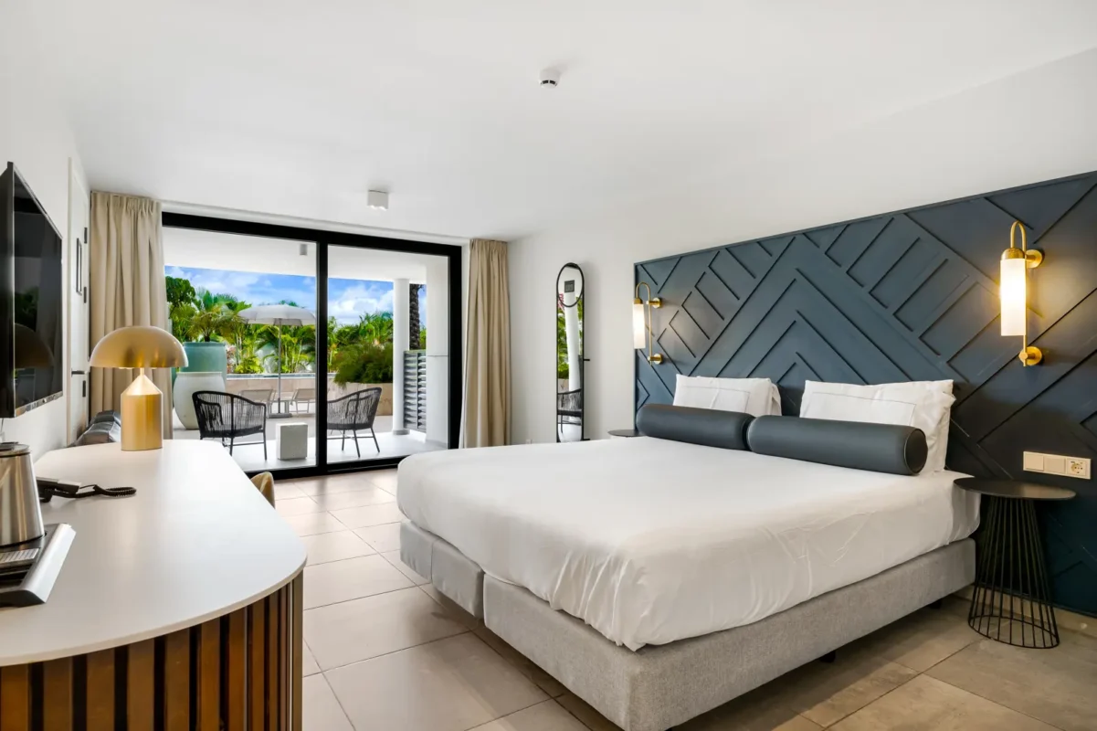 Modern hotel room with a large bed, blue geometric headboard, and balcony doors opening to a tropical outdoor seating area.