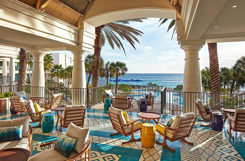 Outdoor resort terrace with lounge chairs and colorful stools overlooking a blue ocean and palm trees.