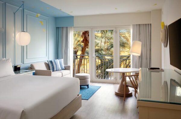 Bright hotel room with a white bed, blue accent wall, seating area, and balcony doors overlooking palm trees outside.