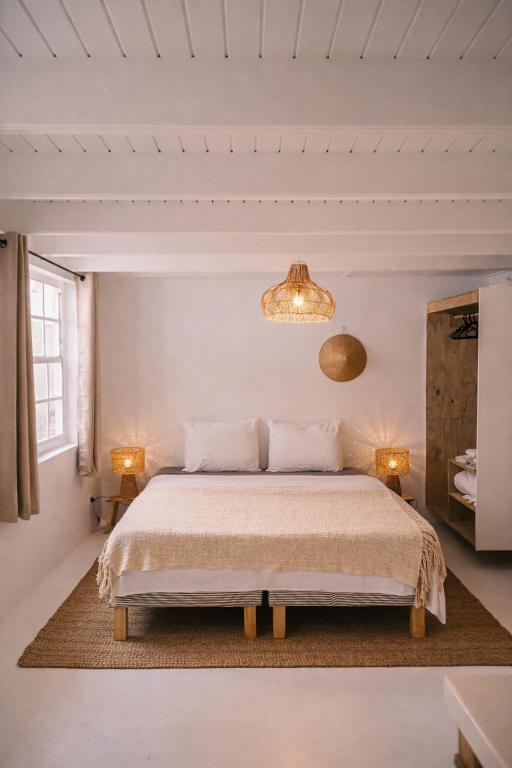 Bright minimalist bedroom with a central bed dressed in white linens, two bedside lamps, a woven pendant light, a beige rug, and an open wardrobe on the right near a window.