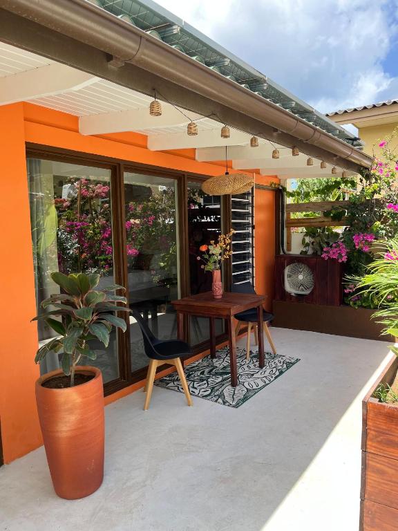 A cozy outdoor balcony with an orange wall, glass doors, and a small wooden table flanked by two chairs, potted plants, and string lights overhead.