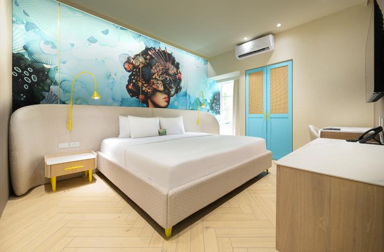 Hotel bedroom with a large white bed, beige headboard, yellow accents, and a mural of a woman underwater on the wall behind it.