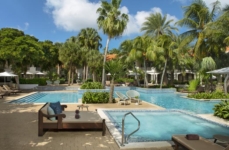 Resort pool area with palm trees, lounge chairs, and a shaded seating area by the water