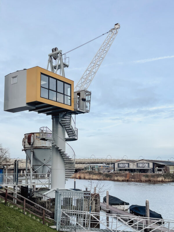 Harbor crane with a yellow cabin on a tall gray tower by a river, with a spiral staircase and docked boats; FoxGym building visible across the water.