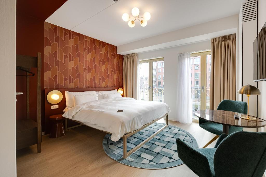 Hotel bedroom with a queen bed against a red geometric accent wall, round blue rug, and bedside lamps by balcony doors.