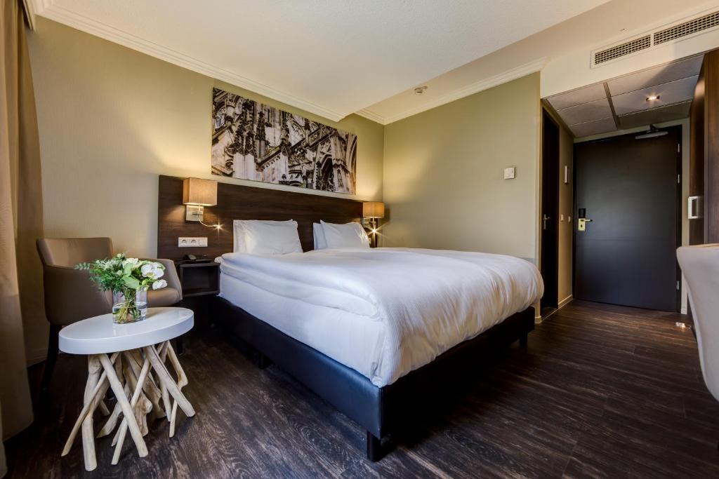 A modern hotel room with a large white‑bed, two nightstands, and wall art above the headboard; a small table with flowers sits by a chair to the left.