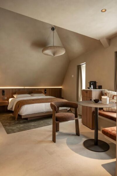 Hotel room with a queen bed, warm wood headboard, and a circular pendant light under a sloped ceiling; a round table with two chairs and a coffee station nearby.