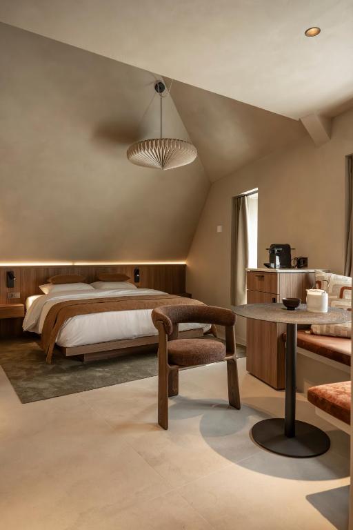Hotel room with a queen bed, warm wood headboard, and a circular pendant light under a sloped ceiling; a round table with two chairs and a coffee station nearby.