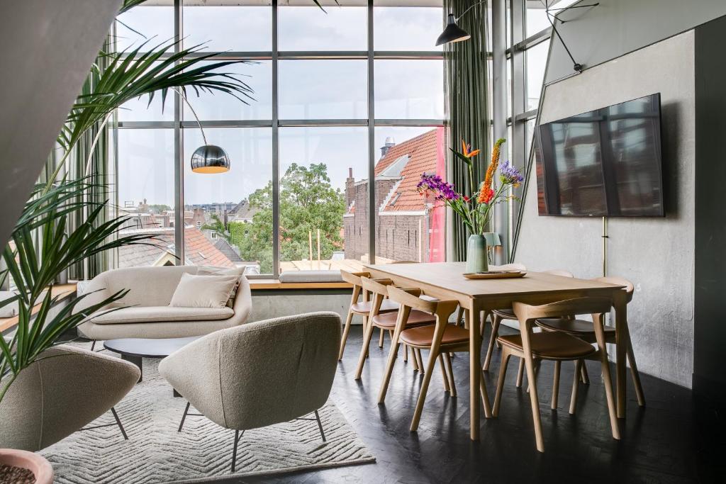 Bright open-plan living and dining area with large windows, beige sofa, two armchairs, and a wooden dining table with a vase of colorful flowers nearby.