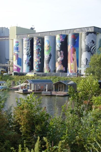Row of tall silos painted with colorful graffiti murals beside a canal, with houseboats on the water in the foreground.