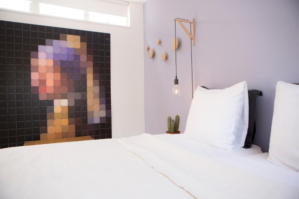 Bedroom with a large pixel-art mosaic on the left wall, white bed, and a hanging bulb light beside it.