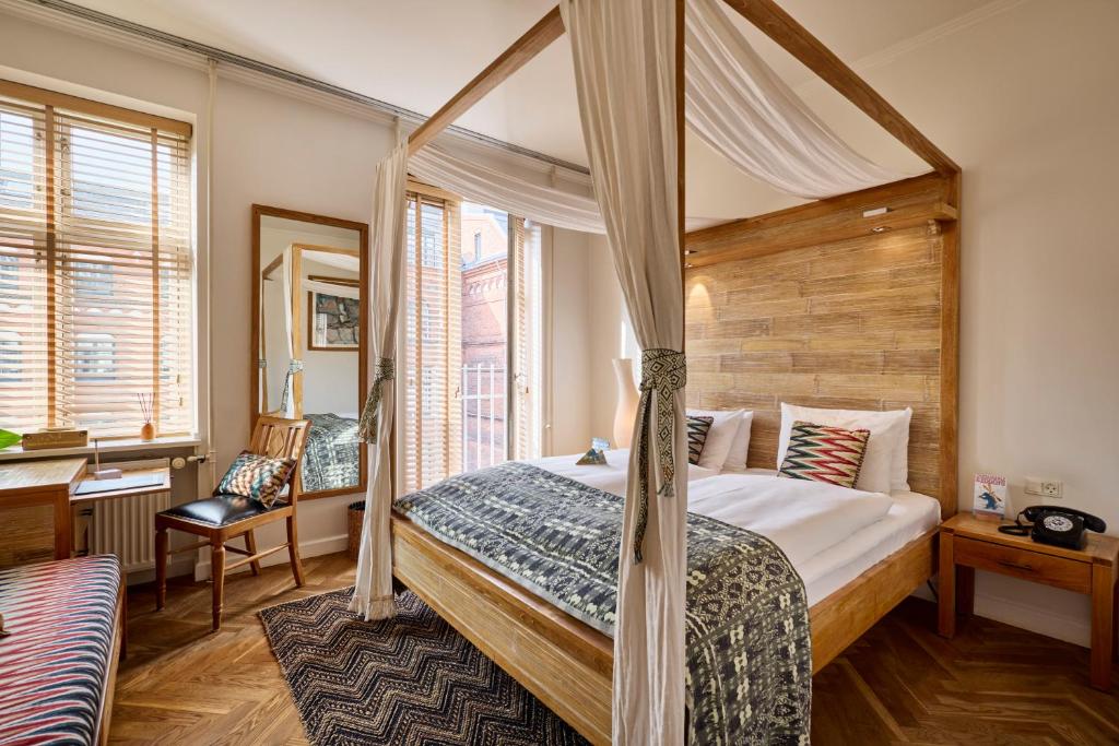 Bright hotel room with a four-poster wooden bed, white linens, and patterned throw; warm natural light from large windows and a woven rug on herringbone floor.