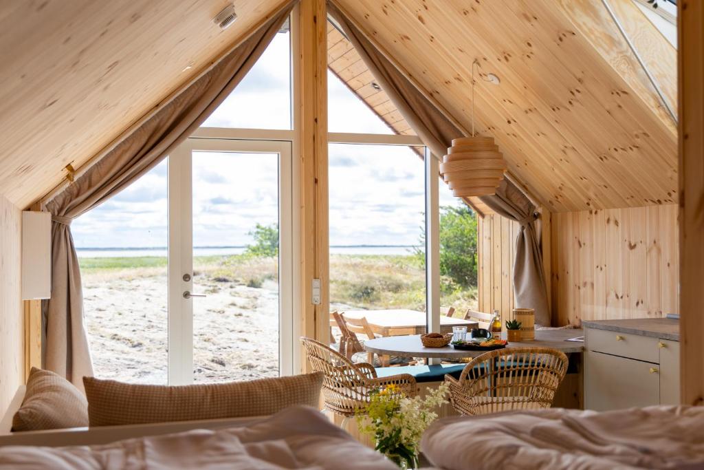 Wooden A-frame interior with a dining table and wicker chairs, opening to a sandy beach and sea through glass doors.