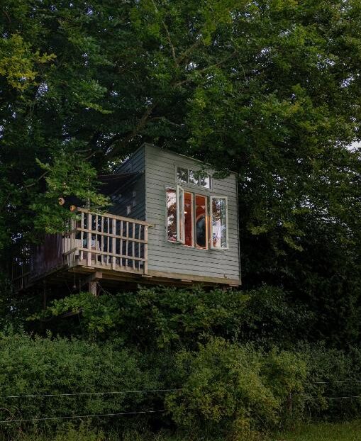Tiny treehouse on stilts with a small deck, nestled among green trees and foliage.