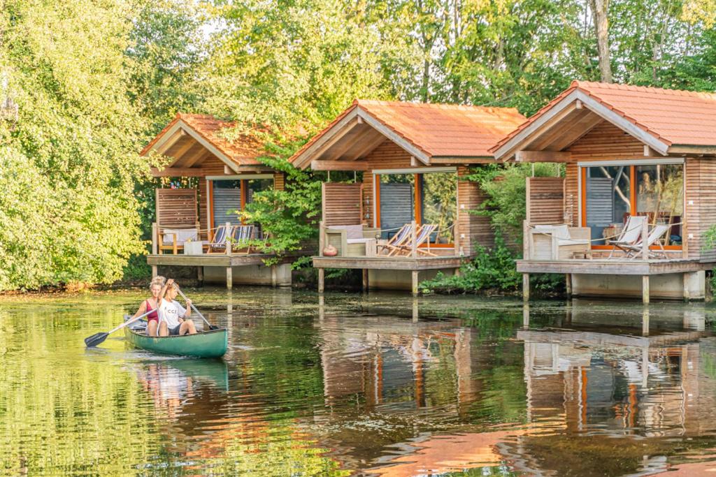 Two people paddle a green canoe on a calm lake in front of wooden bungalow cabins with orange roofs.