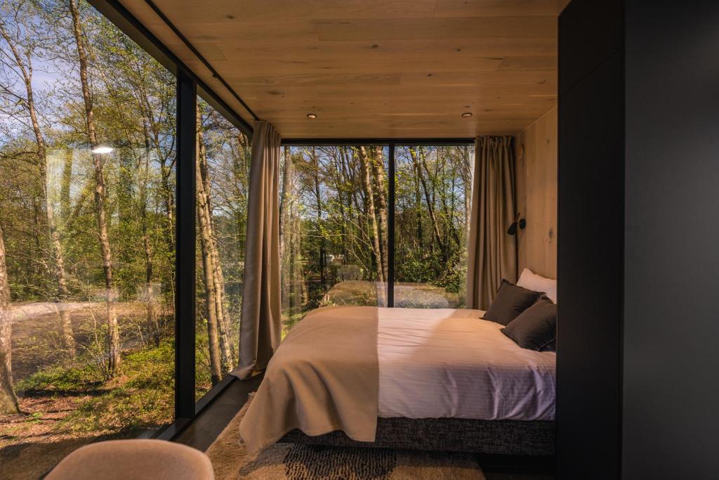 Bedroom with floor-to-ceiling glass walls looking out on a forest, a neatly made bed with white linens and beige throw, and curtains on either side.