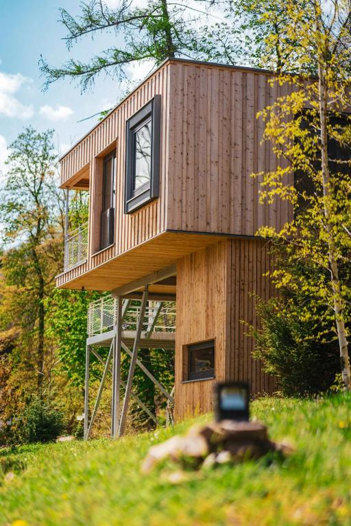 Two-story wooden tiny house on stilts in a sunlit forest, with a deck and large window, among green trees.
