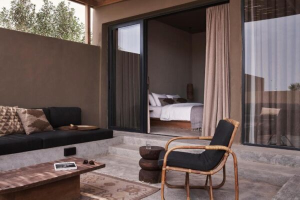 Outdoor patio area with a dark sofa, woven chair, and low wooden coffee table, opening to a bedroom via a sliding glass door with beige curtains.