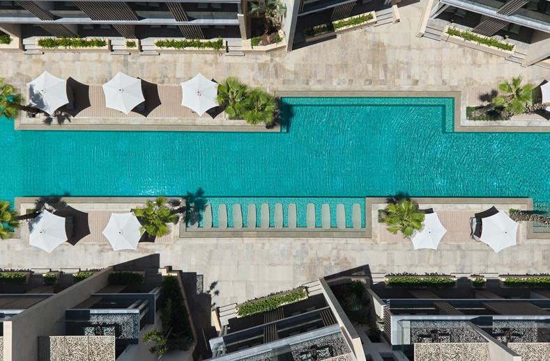 Aerial view of a turquoise rectangular swimming pool flanked by lounge chairs and white umbrellas on a resort deck, with surrounding buildings and palm plants nearby in a top-down shot.